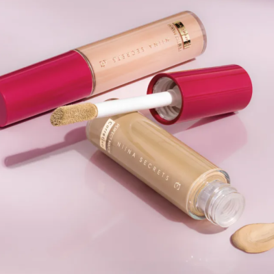 Frascos de corretor líquido Nina Secrets com aplicador aberto e creme amarelado em superfície rosa claro