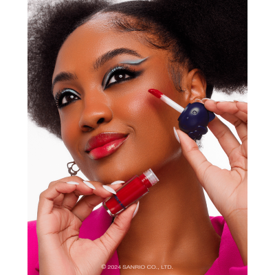 Gloss labial vermelho com tampa azul em mãos de mulher maquilhada com olhos azuis e negros e roupa rosa.