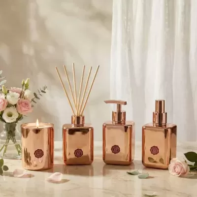 Conjunto decorativo em vidro bronze com vela, difusor, dispenser e spray junto a flores e pétalas numa superfície marmoreada