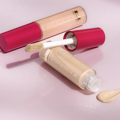 Dois tubos de corretor líquido bege claro com tampa vermelha e aplicador, da marca NINA SECRETS, em fundo branco