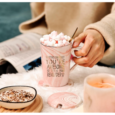 Caneca rosa com marshmallows e texto em inglês ao lado de prato de bolachas e vela rosa