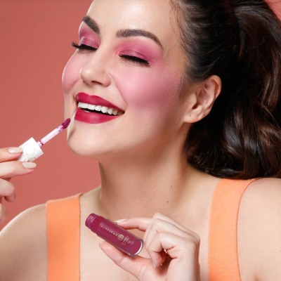 Mulher com maquilhagem rosa a aplicar batom líquido vinho num fundo coral