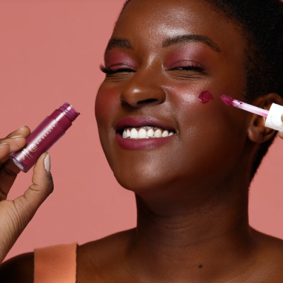 Mulher a aplicar baton líquido rosa-escuro na face com fundo rosa