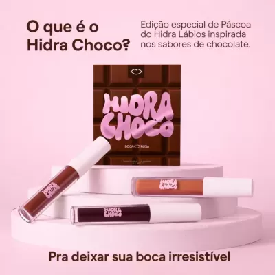 Batons líquidos Hidra Choco com embalagem castanha e texto rosa, sobre suportes brancos e fundo rosa claro.