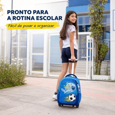 Criança com mochila de rodinhas azul com tema de futebol em ambiente escolar exterior