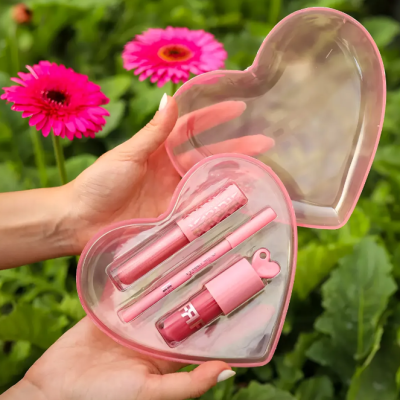 Estojo coração transparente rosa com maquilhagem cor-de-rosa seguro por mãos entre flores em fundo verde
