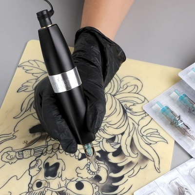 Máquina de tatuagem preta sendo usada sobre uma base com desenho de caveira e flores, agulhas de tatuagem embaladas à parte.