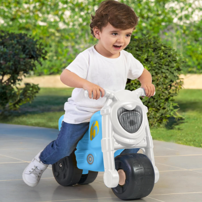 Criança em mota de brincar azul e branca em piso exterior cinzento junto a arbustos