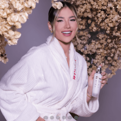 Mulher com roupão branco segurando frasco branco com dispensador, flores secas ao fundo