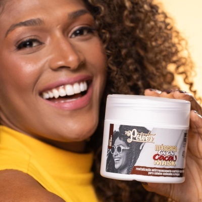 Máscara capilar em frasco branco com rótulo que mostra mulher de óculos e texto promocional, segurado por mulher com cabelo encaracolado e roupa amarela