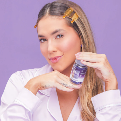 Mulher loira com pijama branco segurando frasco de líquido roxo