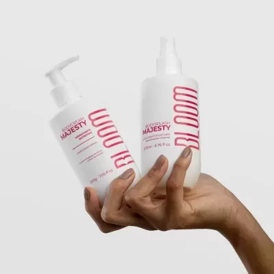 Duas mãos segurando frascos brancos de bodycream e bodysplash Majesty com texto rosa