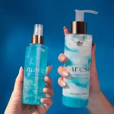 Duas mãos segurando produtos de beleza azul e branco Maresia Toque do Oceano com tampas douradas sobre fundo azul.