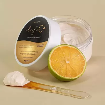 Creme facial iluminador em pote com tampa preta e dourada, colher com creme e meio limão cortado ao lado em fundo bege.