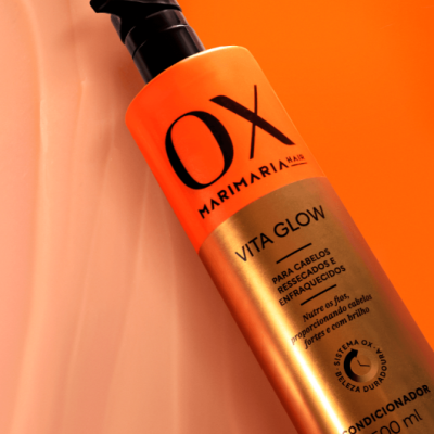 Frasco laranja e dourado de condicionador OX Marimaria Hair sobre fundo laranja e creme
