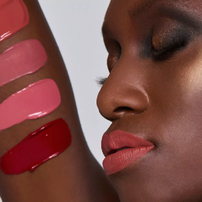 Mulher negra com batom rosa e amostras de batons em tons rosa e vermelho no braço.