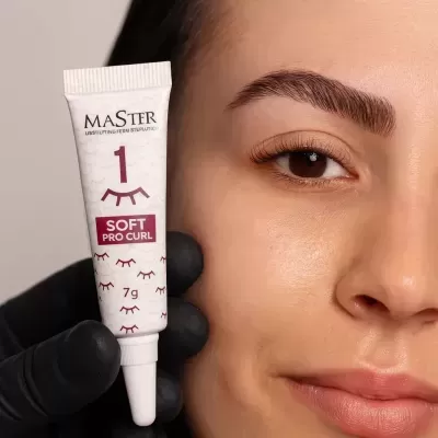 Tubo de cosmético branco segurado por uma mão com luva preta ao lado do rosto de uma mulher com sobrancelhas definidas