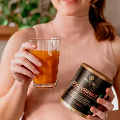 Produto colagénio em pó Essential COLLAGENGUT e copo com bebida laranja nas mãos de mulher com camisola rosa.