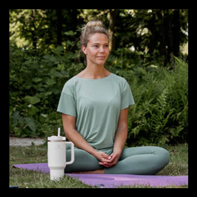 Mulher sentada de pernas cruzadas num tapete de ioga roxo num jardim com roupa desportiva verde-menta e copo térmico branco