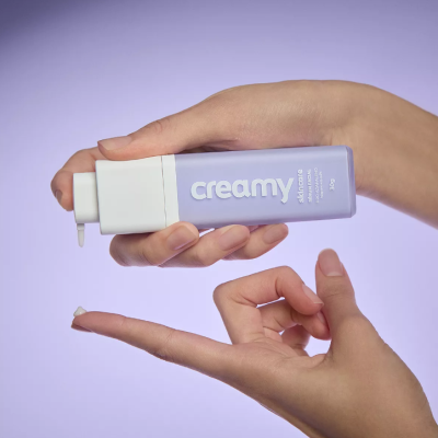 Frasco lilás com tampa branca e texto 'creamy' segurado por mãos femininas