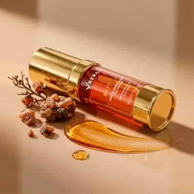 Frasco de óleo facial vermelho e dourado Aberl numa superfície bege com gotas de óleo e ramo seco