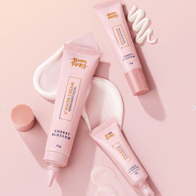 Três tubos de creme hidratante rosa com texto sobre um fundo rosa e cremes espalhados ao redor