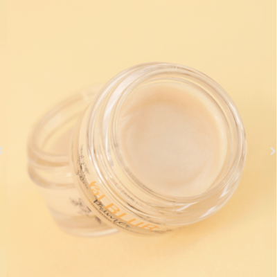 Pote de vidro transparente com pomada bege claro em fundo amarelo