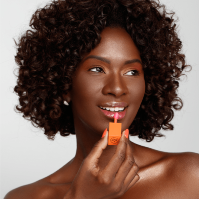 Mulher a aplicar batom rosa com embalagem laranja em fundo cinzento