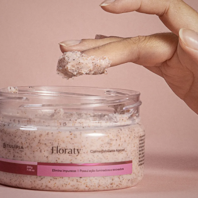 Creme esfoliante floraty em pote transparente com textura rosa granulada e mão a colher produto