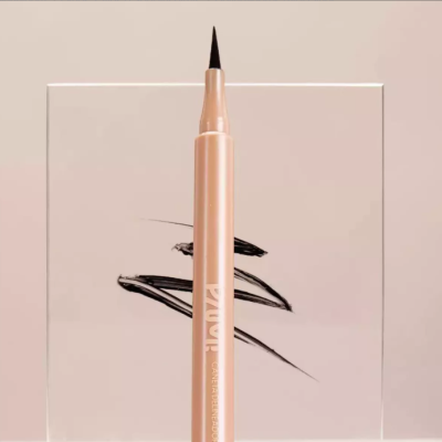 Delineador líquido em caneta bege com ponta preta, texto TAWLA, sobre superfície transparente com traços pretos