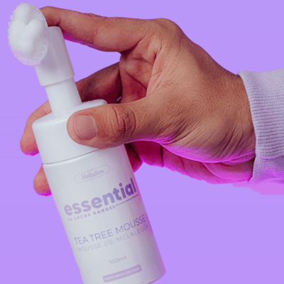 Frasco branco de mousse de Tea Tree segurado por uma mão em fundo roxo