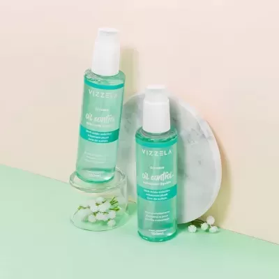 Dois frascos de líquido azul claro da marca VIZZELA com tampas brancas sobre base de vidro e fundo com pedra redonda branca e flores pequenas.