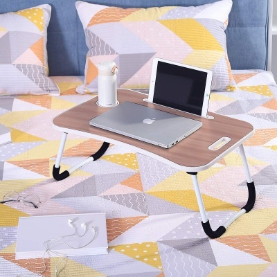 Mesa dobrável com portátil, tablet e garrafa sobre cama com colcha e almofadas coloridas geométricas