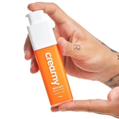 Sérum facial Creamy em frasco laranja com tampa branca, segurado por mãos tatuadas