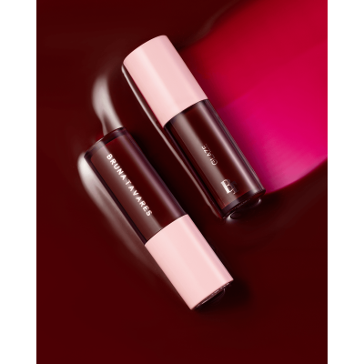 Duas embalagens de batom líquido vermelho escuro com tampas rosa sobre superfície vermelha.