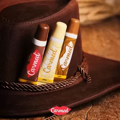 Três batons Carmed em tons castanhos, bege e caramelo sobre chapéu castanho de camurça