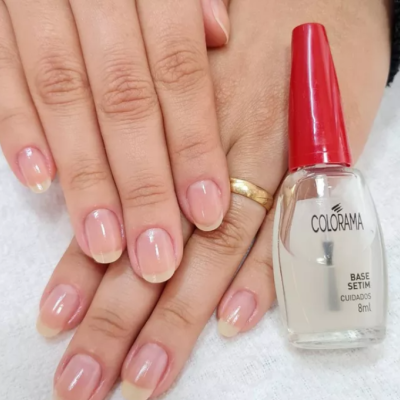 Unhas naturais ao lado de verniz Colorama Base Setim com tampa vermelha sobre tecido branco.