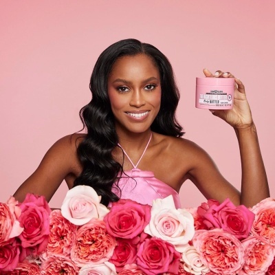 Mulher morena com vestido rosa segurando creme corporal rosa The Righteous Butter entre flores rosas e vermelhas