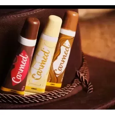 Três sticks de bálsamo labial Carmedi em embalagens vermelha, branca e castanha sobre um chapéu de feltro castanho com corda