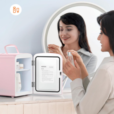 Mini frigo rosa para cosméticos numa mesa junto a espelho redondo com mulher a segurar frasco prateado