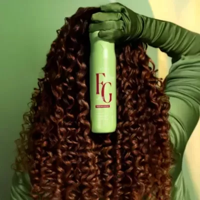 Frasco verde claro com letras vermelhas FG segurado por mão com luva verde sobre cabelo cacheado castanho