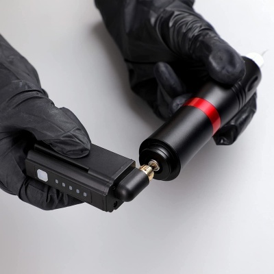 Mãos com luvas pretas segurando máquina de tatuagem preta com faixa vermelha e bateria preta em fundo cinzento