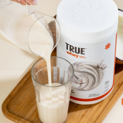 Suplemento alimentar TRUE whey branco e laranja com bebida cremosa a ser servida em copo de vidro.