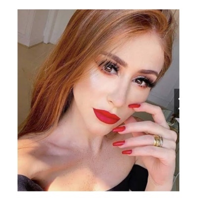 Mulher com cabelo laranja-avermelhado, maquilhagem vermelha, unhas vermelhas, anel dourado e vestido preto