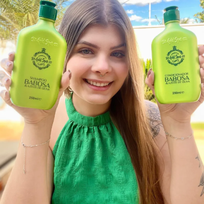 Mulher com blusa verde segurando frascos verdes de shampoo e condicionador Babosa ao ar livre