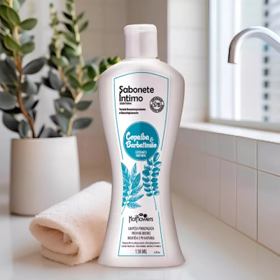 Frasco branco de sabonete íntimo com texto azul e preto, ao lado de toalha branca e planta em vaso branco na bancada.