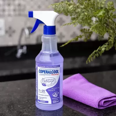Frasco de spray roxo com tampa branca e azul ao lado de pano de microfibra roxo sobre bancada escura com planta verde ao fundo.