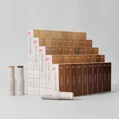 Embalagens e sticks de maquilhagem Boca Rosa em vários tons de bege, castanho e roxo sobre fundo cinza claro.