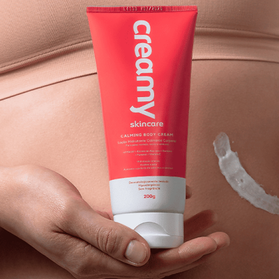 Frasco vermelho e branco de creme corporal creamy skincare segurado por uma mão com creme aplicado na pele ao fundo.