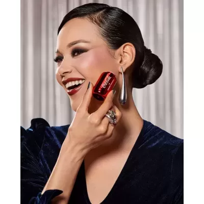 Mulher sorridente com vestido azul e brincos prateados a segurar frasco vermelho COCA-COLA ZERO SUGAR.